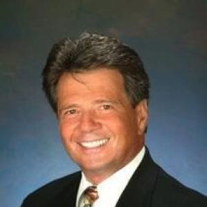 older white man headshot smiling wearing a brown suite white shirt and brown and blue tie