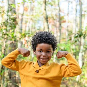 smiling young black boy standing in front of trees wearing a gold sweater and smiling and making muscle man arms