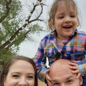 young white couple with their daughter outside little girl has on plaid shirt dad is bald with a mustache and beard mom has long brown hair