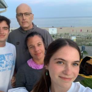 A white family of four the man is bald with glasses wearing a gray hoodie and holding a yellow hat
