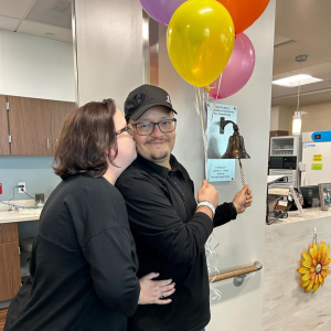 Hispanic couple both dressed in black se is kissing him as he rings the bell and holds balloons