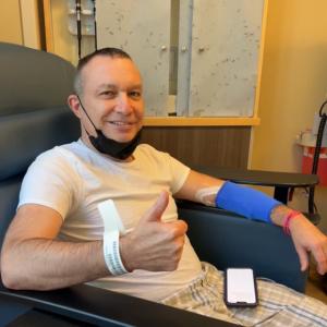 middle age white man with short hair, a black mask pulled down, a blue sleeve on his arm giving a thumbs up wearing white shirt with plaid shorts