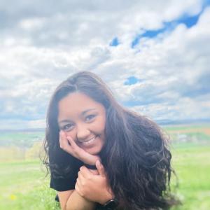 smiling young hispanic girl with long dark hair lying in the grass head propped on hand wearing black shirt and watch