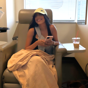 young ethnic girl sitting in a hospital chair wearing a yellow ball cap and a black sleevelss tshirt holding a pink phone with a blanket on her lap