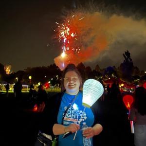 young white girl with her hair pulled up with glasses wearing a black jacket and blue tshirt carrying a white balloon at LTN with fireworks in the background