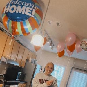 young white woman bald with a nose ring wearing a sweatshirt and standing in a kitchen with balloons and a bundt cake