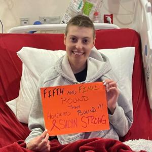 young white woman in a hospital bed with short hair wearing a gray hoodie and holding an orange sign about fifth round of chemo