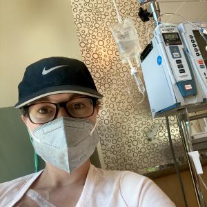 young white woman with short hair wearing a blue ball cap, brown glasses, a face mask and a pink Underarmor shirt