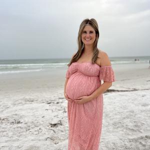 Holly young white woman with long hair and very pregnant wearing a pink off the should lace dress standing on a beach