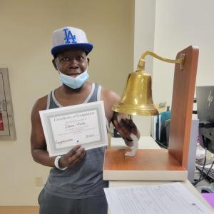 middle aged black man wearing LA ballcap and gray and white tank top and gray sweat pants holding a certificate and ringing a bell.