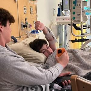 young white teen boy with dark hair lying in a hospital bed with his arm extended over his head looking at an orange phone being held by another teen boy