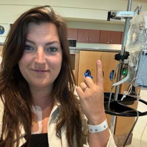 young white woman with long brown hair wearing a black shirt and white jacket sitting in a hospital room holding up her pointer finger