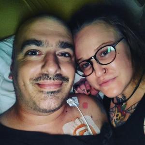 younger white man with bushy eyebrows and mustache wearing a black shirt with a port lying next young white woman with black glasses and a black nose ring with a tattoo on her finger and neck