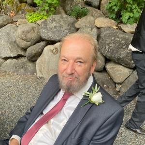 Gary older white man with blue eyes and thinning hair and gray beard and mustache leaning on rocks wearing a blue suite, mauve tie and a rose boutonaire
