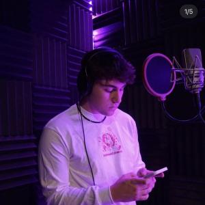 Marko young white male with curly dark hair in a recording studio wearing headphones and a puka shell neclklace and a white long sleeve tshirt with a pink logo also holding a phone