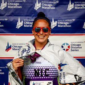 Nicolas smiling middle aged hispanic woman with dark hair pulled up in a bun wearing orangish sunglasses and a Team In Training Shirt holding a medallion in front of a Chicago Marathon banner.jpg