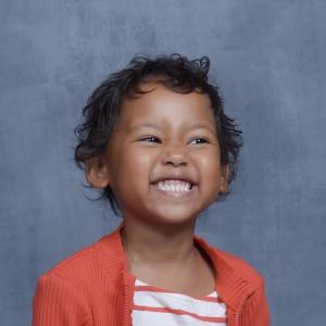Sofia school picture of pre-K black girl with short curly hair and a big smile wearing and orange and white striped top under an orange sweater with a zipper
