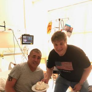 Greg two middle aged white men, one sitting on a hospital bed wearing a grey t-shirt and black shorts the other wearing a flag shirt and jeans both holding a cake
