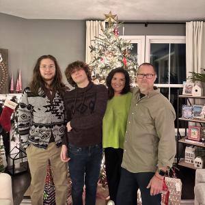 John William middle aged white man wearing glasses and a gray shirt standing in front of Christmas tree with his wife in a green sweater and two sons
