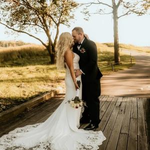 Kayla young woman with long blond hair in a white spagetti strap wedding dress holding a bouquet with arm around a young white man with beard and mustache.