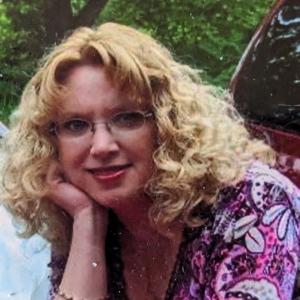 middle aged white woman with curly blonde hair and glasses with her chin resting on her hand wearing a pink and purple print shirt