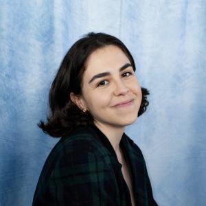 Crystal young hispanic woman with dark hair and gold hoop earrings smiling and wearing a blue and plaid top with blue photo background