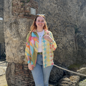 Eva young white woman with reddish hair sunglasses on her head in a white tshirt under a multi color shirt with blue jeans standing in front of a stone wall.