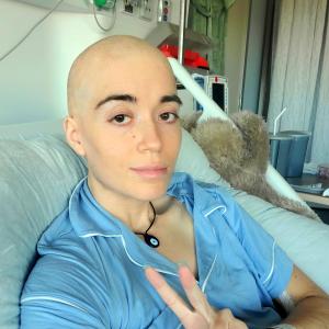 Josie young white woman bald with an orange tatoo and wearing blue pajamas in a hospital bed giving the peace sign