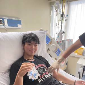 Kevin young asian young man with black hair wearing a puka shell necklace and black gym tshirt and shorts while laying in a hospital bed
