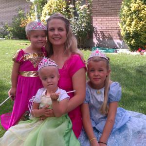 Kim young white mother with long blond hair in a pink dress with three little girls wearing princess outfits and crowns sitting outside
