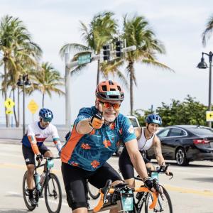 Michael older white man with white beard and mustache wearing an orange bike helmet and blue and orange shirt riding a bike while giving a thumbs up