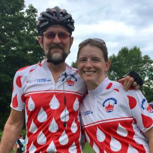 Paul older white gentleman with beard and mustache wearing a bike helmet and sunglasses with younger white female with freckles and sunglasses on her head both wearing Scenic Shore 150 shirts