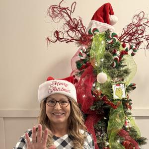 Lori middle aged white woman with long blond hair wearing a Merry Christmas santa hat and glasses and black and white geometric blouse standing in front of a Christmas tree