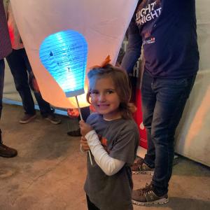 young smiling white girl in a gray shirt over a white long sleeved shirt with and orange bow in her hair and holding a LTN balloon