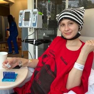 young white woman wearing a black and white knit cap and a red t-shirt and black short with painted white fingernails laying in an infusion chair