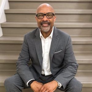 Arturo Pierre middle aged black man bald with mustache and beard wearing black glasses and a grey suite and white shirt sitting on steps