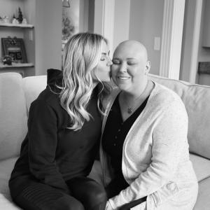 Janie young white woman who is bald wearing a black shirt and a white speckled cardigan being kissed by a young white woman with long blond hair wearing a black hoodie both sitting on a couch