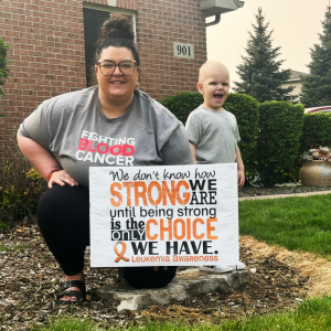 Maverick young white mother with a bun and glasses wearing gray blood cancer shirt holding a leukemia sign with her young son next to her in a gray shirt and smiling