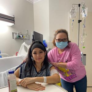 Mayra young hispanci woman sitting in a hospital bed wearing a black headbank and blue hospital gown and has three face piercings with another woman with face mask and pink shirt next to her