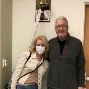 Rex older white couple standing in a hospital room man has white hair and glasses wearing a gray jacket and pajama pants woman has face mask