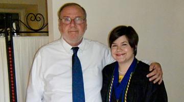 Abby, right, a young white woman with short hair, stands in a graduation gown next to her father, an older white man wearing a shirt and tie.