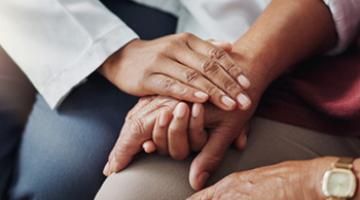 A person with dark-skinned hands clasps the hand of a second dark-skinned person to offer comfort.