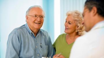 An older, white man smiles and is joined by his wife, who is also white, as he speaks to his doctor