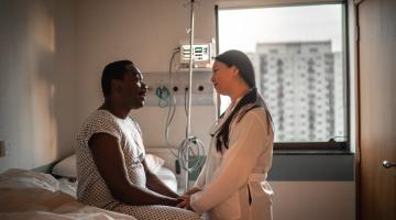An African-American patient holds the hand and speaks with a doctor of Asian descent in a hospital room