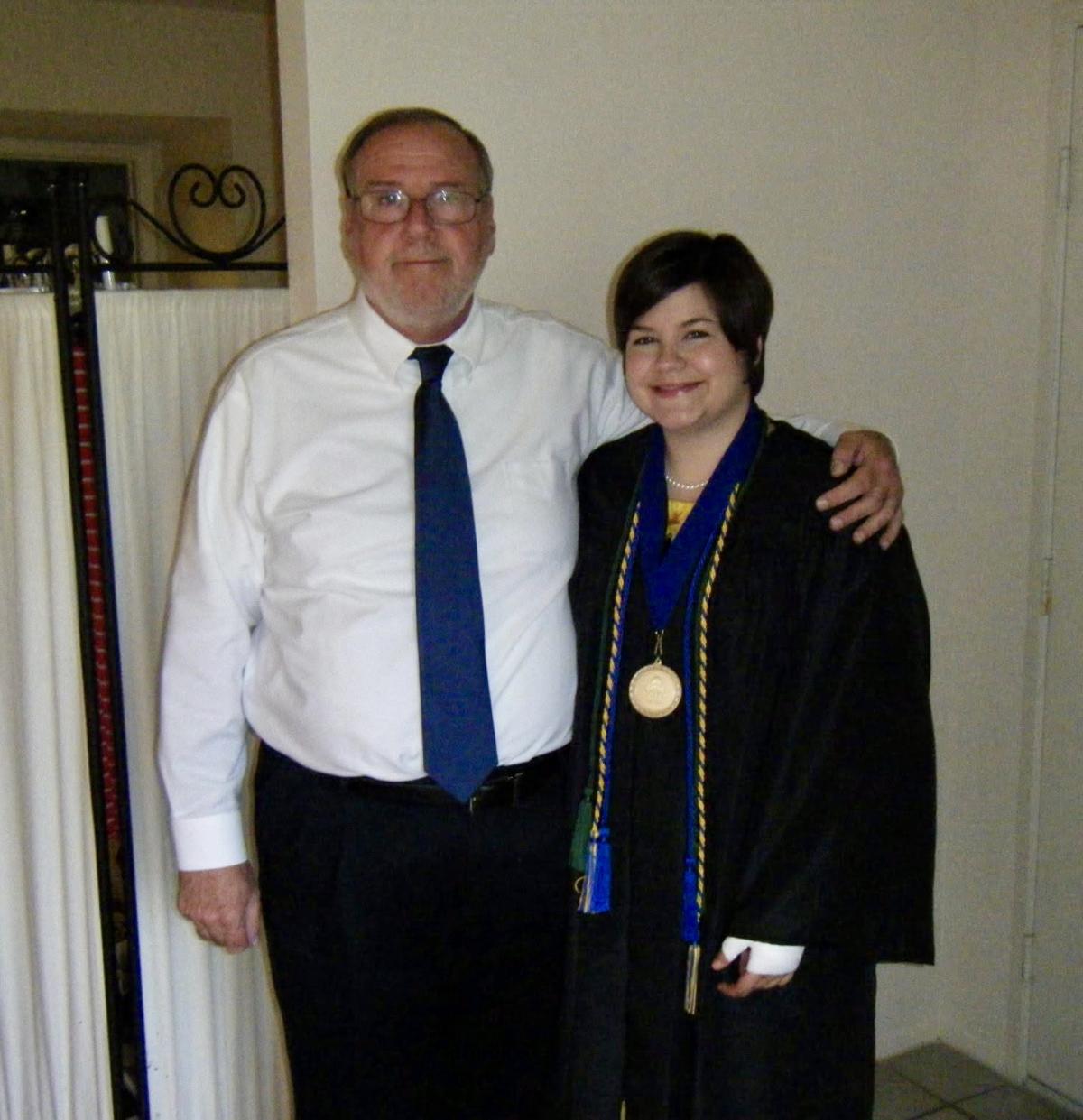 Abby, right, a young white woman with short hair, stands in a graduation gown next to her father, an older white man wearing a shirt and tie.