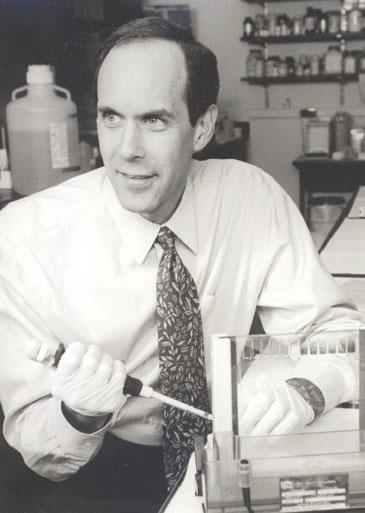 Brian Druker, a white man, wears a button-down shirt and tie and holds a medical pipette in a research laboratory.