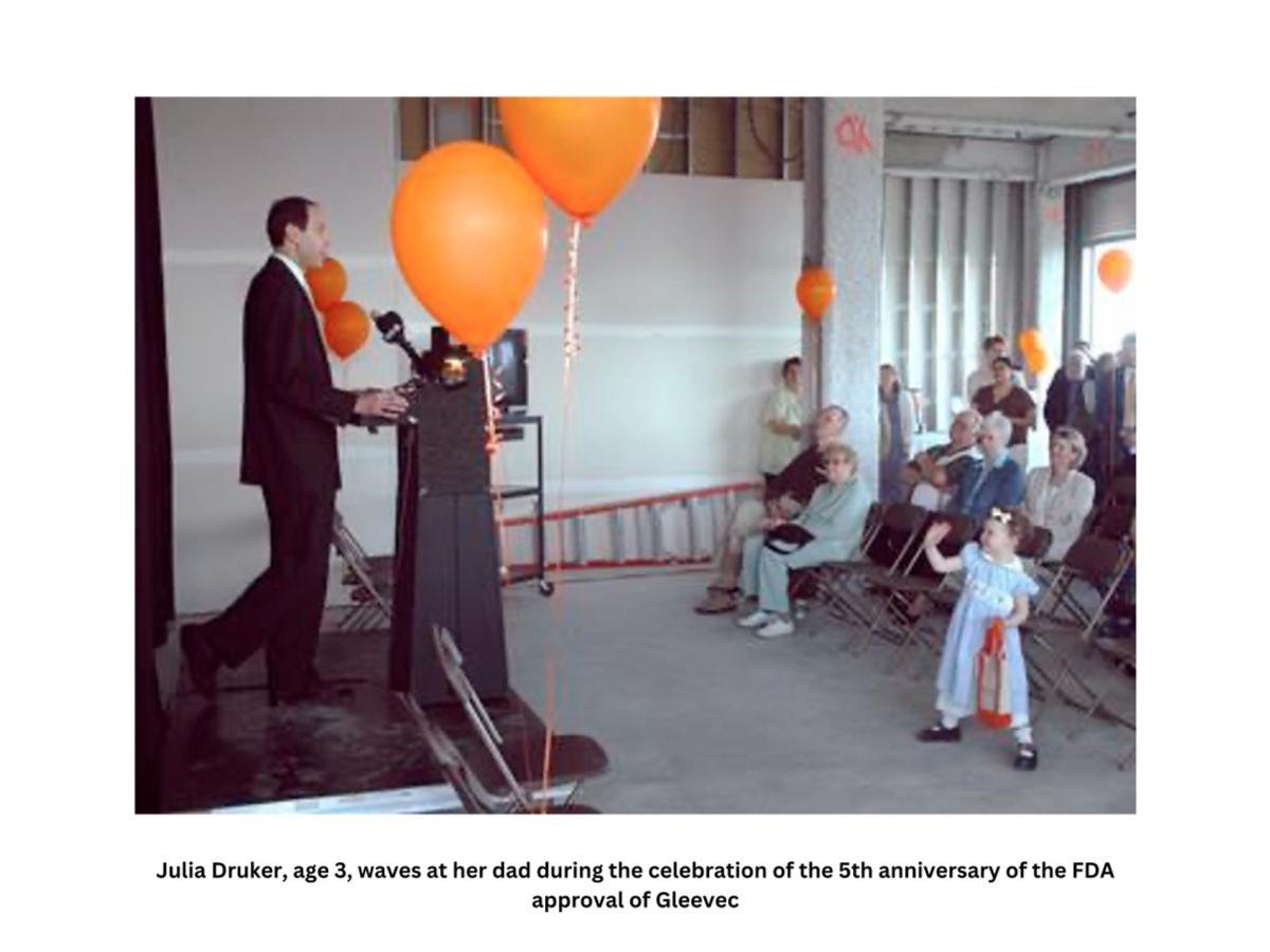 A man stands at a speaking podium, surrounded by orange balloons. He's facing a small crowd of people sitting in folding chairs or standing as they listen to him. A young girl wearing a dress and hair bow waves in the foreground.