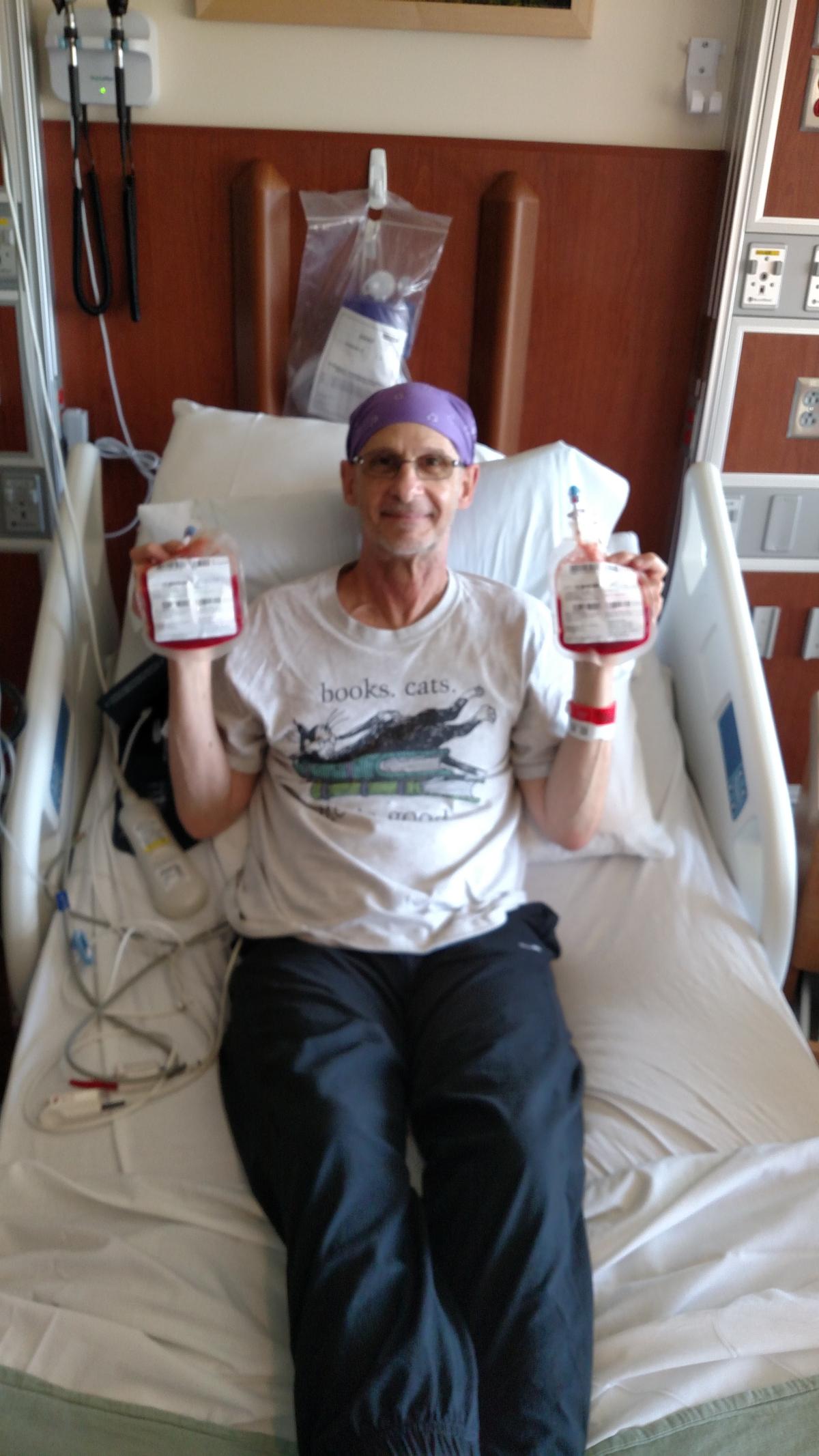 Steve is a white man with glasses, wearing a purple bandana, a white shirt, and black pants. He sits in a hospital bed, holding two medical bags bearing the names of his bone marrow donors.