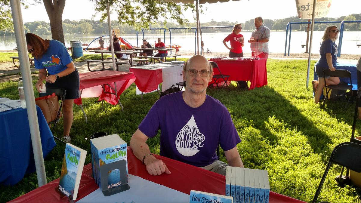 Steve, a white man with glasses, sits behind a table at an event in a park, with stacks of books on the table next to him.