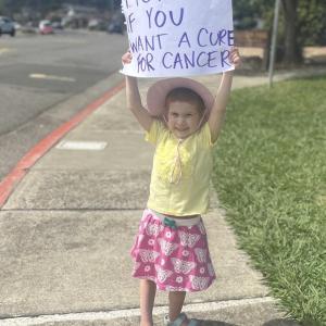 little white girl in a yellow shirt and pink skit with butterflies on it wearing a hat and holding up a sign that says honk if you want a cure for cancer